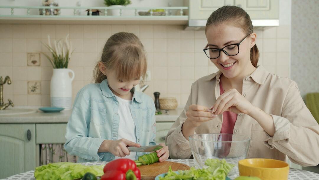 Helpful daughter is making vegetable salad chopping cucumber while young loving mother is touching her hair expressing love at home in kitchen.